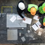 Workers and manager in safety helmets working with documents at factory table